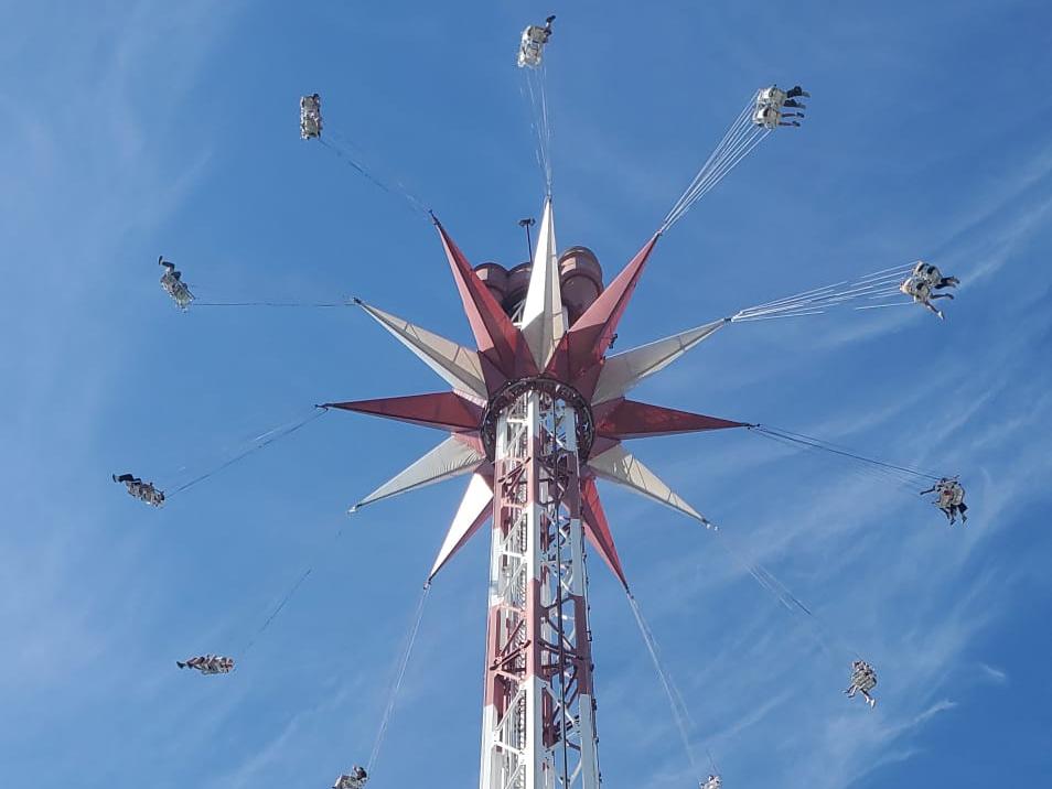 Ein hohes Kettenkarussell ragt vor einem strahlend blauen Himmel in die Höhe. Die Fahrgäste schwingen in ihren Sitzen an langen Ketten kreisförmig um den rot-weiß gestalteten Turm. Die Aufnahme wurde aus der Untersicht gemacht und betont die Höhe der Attraktion
